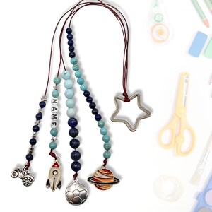 Może przedstawiać: Zestaw czterech kolorowych zakładek z koralików z różnymi zawieszkami, w tym rakietą, planetą, piłką nożną i traktorem. Koraliki są niebieskie, turkusowe i białe. Zakładka jest przymocowana do czerwonej sznurówki i ma srebrny zawieszkę w kształcie gwiazdy na górze.