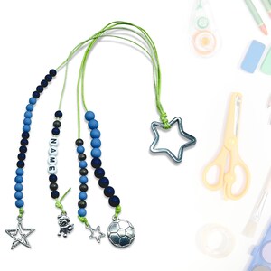 Może przedstawiać: Trzy niebiesko-czarne zakładki z koralików z różnymi zawieszkami. Zawieszki to gwiazda, piłka nożna i kreskówkowa postać. Zakładki są nawleczone na zieloną sznurówkę.