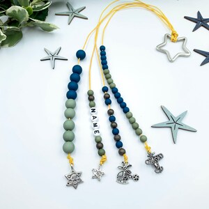 Może przedstawiać: Trzy koraliki naszyjniki z różnymi wisiorkami. Pierwszy naszyjnik ma wisiorkiem w kształcie gwiazdy, drugi ma wisiorkiem w kształcie gwiazdy i słowo "NAME" napisane białymi koralikami, a trzeci ma wisiorkiem w kształcie helikoptera. Koraliki są zielone, niebieskie i złote.