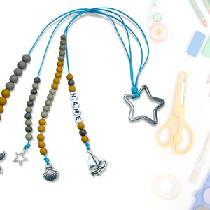 Może przedstawiać: Zestaw trzech zakładek z koralikami i różnymi zawieszkami. Koraliki są brązowe i szare. Zawieszki to gwiazda morska, muszla i żaglówka. Zakładki są połączone niebieską wstążką. Na jednej z zakładek napisane jest "NAME".