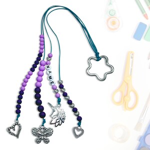 Może przedstawiać: Fiorek z koralików w kolorze fioletowym i turkusowym z zawieszką w kształcie gwiazdki, zawieszką w kształcie jednorożca, zawieszką w kształcie motyla i dwoma zawieszkami w kształcie serca. Fiorek ma również metkę z imieniem, na której widnieje napis "NAME".