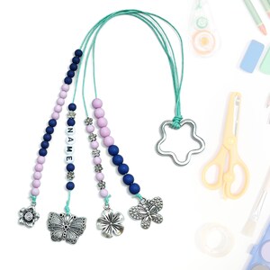 Może przedstawiać: Turkusowy sznurek z różowymi, niebieskimi i przezroczystymi koralikami. Sznurek ma srebrny zawieszkę w kształcie gwiazdy, srebrny zawieszkę w kształcie kwiatu i srebrny zawieszkę w kształcie motyla. Koraliki tworzą słowo "NAME".