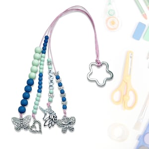 Może przedstawiać: Różowo-biały sznurek z koralikami z zawieszką w kształcie gwiazdy, niebieskimi i zielonymi koralikami oraz srebrnymi zawieszkami, w tym motylami, jednorożcem i sercem. Sznurek ma wyhaftowane słowo "NAME" białymi koralikami.