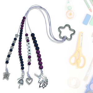 Może przedstawiać: Sznur z koralików w kolorze fioletowym i czarnym z zawieszką w kształcie srebrnej gwiazdy, zawieszką w kształcie motyla, zawieszką w kształcie baletnicy, zawieszką w kształcie serca i zawieszką w kształcie jednorożca. Sznur ma również koraliki, które tworzą słowo "NAME".