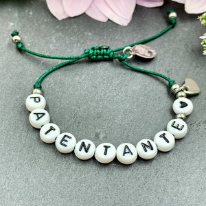 Op de afbeelding: Een groen koordarmband met witte kralen die "PATENTE ANTE" spellen en een zilveren hartbedel.