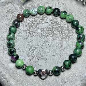 Op de afbeelding: Een armband van groene en zwarte edelstenen met een zilveren hartvormige hanger. De armband is gemaakt van ronde kralen en is geregen op een zilveren ketting.