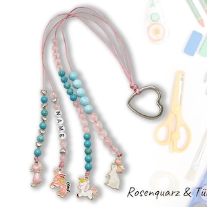 Może przedstawiać: Trzy różowe i turkusowe naszyjniki z koralików z zawieszkami. Zawieszki to kot, jednorożec i królik. Naszyjniki są nawleczone na srebrny wisiorek w kształcie serca, a na jednym z naszyjników napisane jest "NAME". Naszyjniki leżą na białym tle z przyborami szkolnymi.
