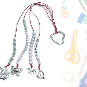 Może przedstawiać: Trzy różowe i szare koraliki z zawieszkami. Zawieszki to jednorożec, motyl i serce. Naszyjniki są przymocowane do srebrnego pierścienia w kształcie serca.