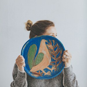 May include: A woman is holding a blue ceramic plate with a painted design of a pink bird with green and brown accents.