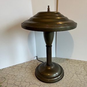 May include: An antique brass desk lamp with a round, tiered shade and a fluted column base. The lamp has a small decorative finial on top and a circular base. The cord is visible, suggesting it is in working condition.