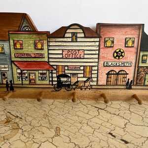 May include: Wooden wall-mounted key holder featuring a painted western town scene with a general store, post office, and blacksmith shop. The key holder has four wooden pegs and is painted in various colors.
