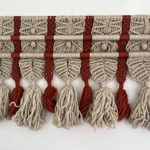 May include: A macrame wall hanging with a beige and rust color scheme. The hanging features a geometric pattern with tassels at the bottom.
