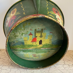 Könnte beinhalten: Ein vintage grüner Metallbehälter mit einer bemalten Szene einer gelben Burg mit rotem Dach und blauem Himmel. Der Behälter hat ein florales Design am Rand.