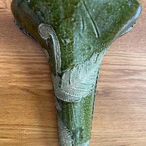 Puede incluir: Un jarrón de pared de cerámica verde con una parte superior acampanada y un cuerpo largo y cónico. El jarrón presenta diseños de hojas de helecho en relieve y un acabado brillante. El jarrón mide unos 25 cm de alto.