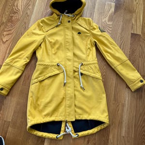 Könnte beinhalten: Gelbe Kapuzenjacke mit schwarzem Innenfutter. Die Jacke hat einen Kordelzug in der Taille, zwei Vordertaschen und ein kleines Regenschirm-Logo auf der Brust. Die Jackenlänge beträgt ca. 76 cm.