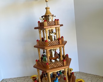 May include: A multi-tiered wooden Christmas pyramid featuring a rotating top fan with snowflake patterns. The structure has four levels with small painted wooden figurines, including angels and animals, and six gold-colored candle holders.