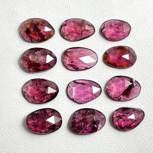 May include: A collection of twelve faceted, oval-shaped pink tourmaline gemstones arranged in a four-by-three grid. Each stone displays a translucent, deep magenta hue with visible internal inclusions and a polished, multi-faceted surface.
