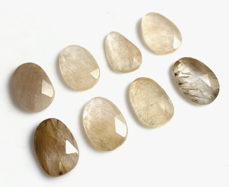 May include: Eight smooth, oval-shaped, light brown and white gemstone cabochons with a faceted edge.