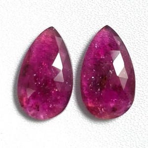 Può includere: Due pietre preziose sfaccettate a forma di pera di un colore magenta-rosa intenso. Ogni pietra presenta una parte superiore appuntita e una base arrotondata, con una superficie lucida che riflette la luce attraverso le molteplici facce.