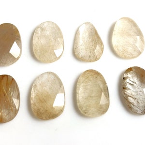 May include: Eight faceted, oval-shaped, translucent, light brown, and gold gemstone cabochons. The stones have a cloudy, milky appearance with gold inclusions.