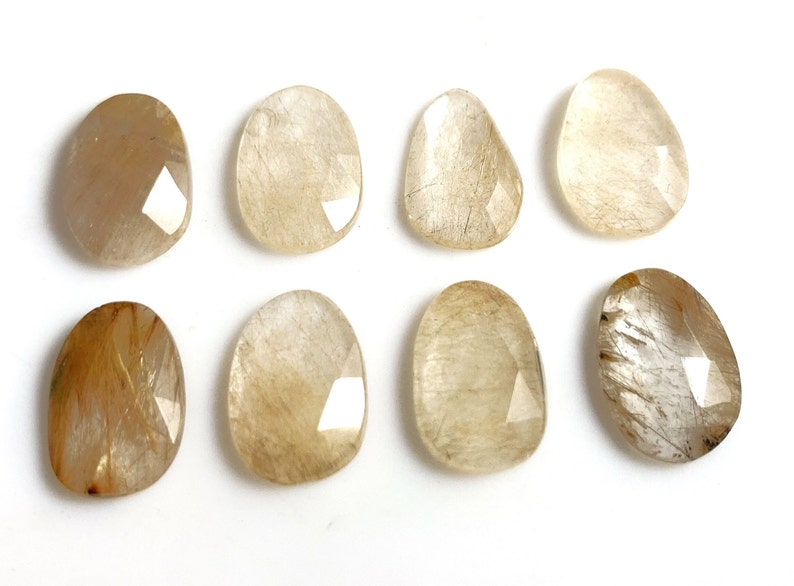 May include: Eight faceted, oval-shaped, translucent, light brown, and gold gemstone cabochons. The stones have a cloudy, milky appearance with gold inclusions.