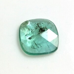 Könnte beinhalten: Ein facettierter, quadratisch geschliffener Edelstein in Türkis. Der Edelstein weist Einschlüsse und eine polierte Oberfläche auf, die das Licht reflektiert. Der Stein ist vor einem weißen Hintergrund platziert, um seine Farbe und Klarheit hervorzuheben. Geeignet für die Schmuckherstellung.