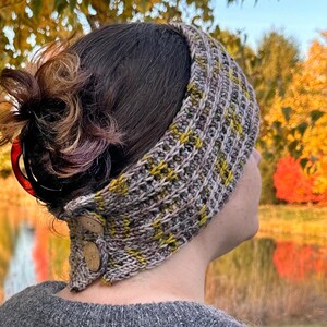 Puede incluir: Diadema de punto en tonos grises, marrones y amarillos, con un patrón texturizado. La diadema tiene dos botones de madera en el lateral. El fondo muestra follaje otoñal y una masa de agua.