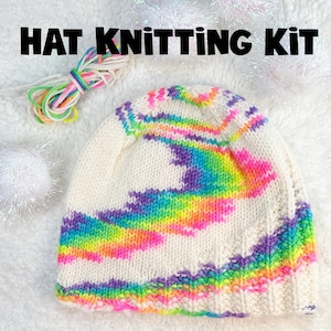 May include: A hat knitting kit with a white knitted hat displaying a rainbow design. The hat has a colourful pattern of pink, orange, yellow, green, blue, and purple. The kit includes a bundle of colourful yarn. Text on the image reads "HAT KNITTING KIT."