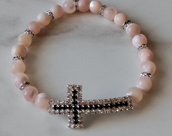 Könnte beinhalten: Ein rosa-weißes Perlenarmband mit einem silbernen Kreuzanhänger und einem Schmetterlingsanhänger mit der Aufschrift "Made with Love".