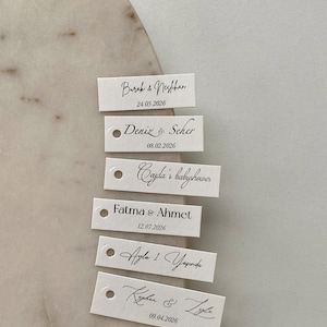 Könnte beinhalten: Eine Sammlung kleiner, rechteckiger weißer Anhänger mit schwarzer Kalligraphie. Jeder Anhänger zeigt Namen und Daten, wahrscheinlich für ein besonderes Ereignis. Die Anhänger sind auf einer hellen, marmorierten Oberfläche angeordnet.