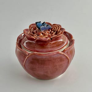 May include: A ceramic jar and lid, designed to resemble a flower in bloom. The jar is a deep reddish-pink with a glossy finish. The lid is adorned with a blue butterfly detail. The jar is a decorative item, suitable for display.