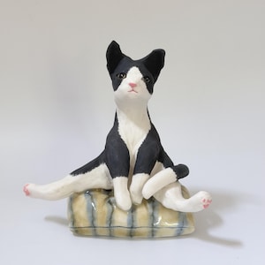 May include: A ceramic figurine of a black and white cat sitting on a rectangular, striped beige and blue cushion. The cat has a white face, chest, and paws, with black ears and back, and small pink details on the paw pads and nose.