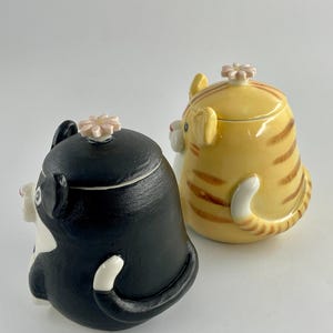 May include: Two ceramic cat-shaped containers. One is black and white, the other is yellow with brown stripes. Both have a flower-shaped lid and a curled tail. The containers are designed to resemble sitting cats.