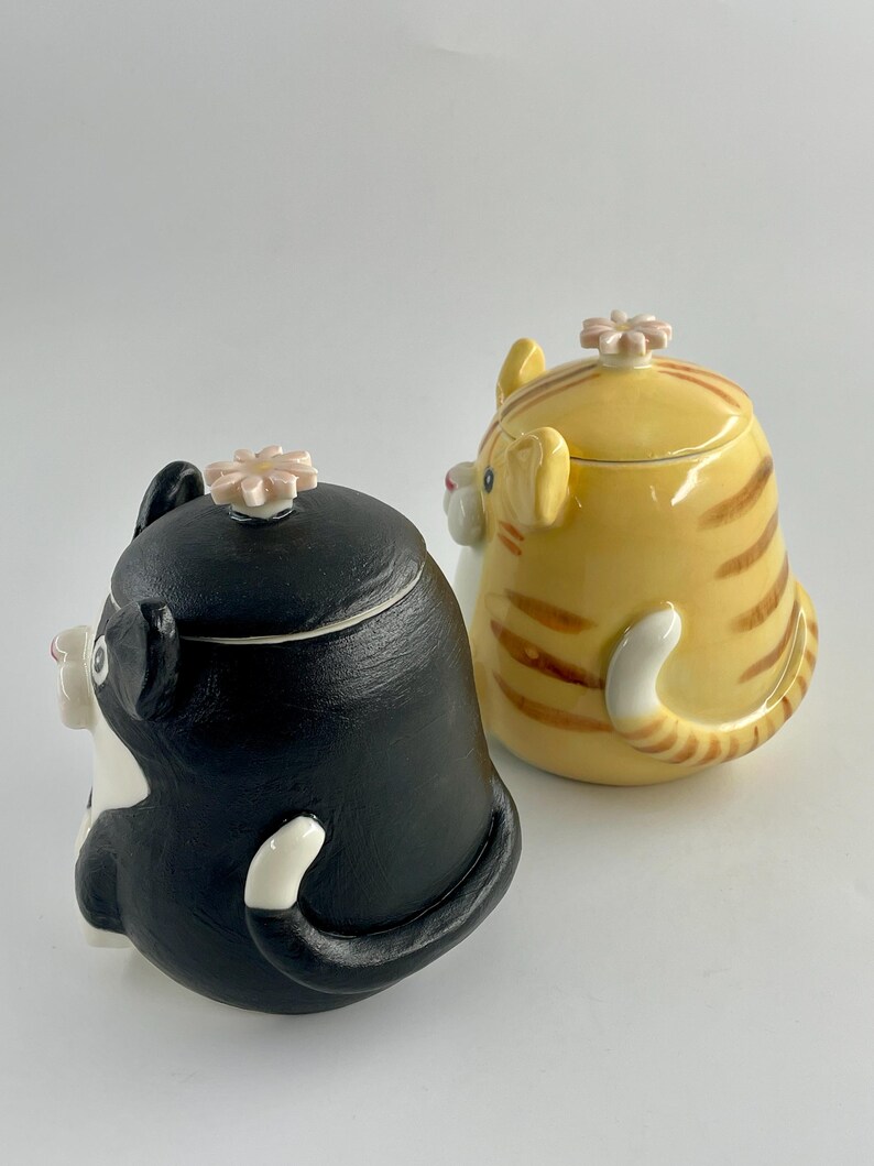 May include: Two ceramic cat-shaped containers. One is black and white, the other is yellow with brown stripes. Both have a flower-shaped lid and a curled tail. The containers are designed to resemble sitting cats.