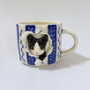 May include: A ceramic mug featuring a white base with vertical blue stripes and a repeating leaf pattern. A three-dimensional black and white cat head emerges from a sculpted white opening on the front, with one paw extending outward.