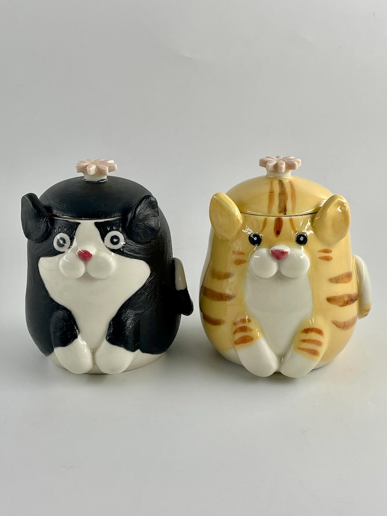 May include: Two ceramic cat-shaped jars. One is black and white, the other is yellow with brown stripes. Both have a pink flower-shaped knob on the lid and white paws. The jars have a cartoonish design.