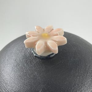 May include: A black ceramic lid with a flower-shaped knob. The knob is light pink with a yellow centre. The lid has a textured surface. The flower knob is attached to the lid with a silver ring.