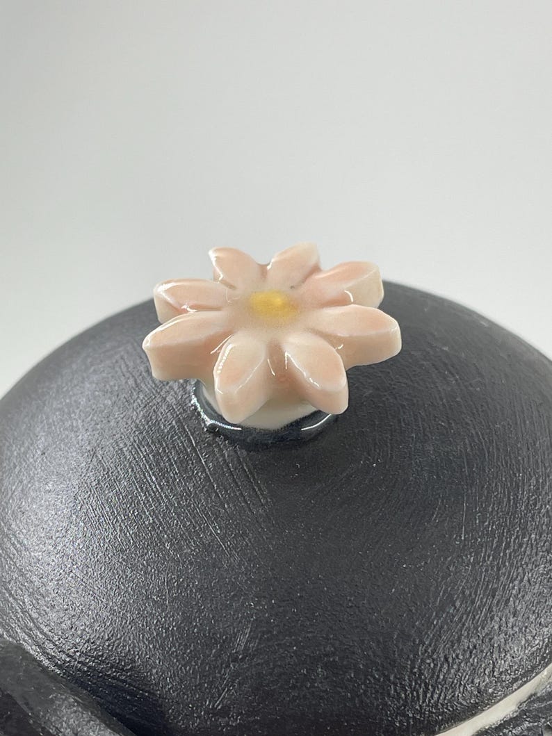 May include: A black ceramic lid with a flower-shaped knob. The knob is light pink with a yellow centre. The lid has a textured surface. The flower knob is attached to the lid with a silver ring.