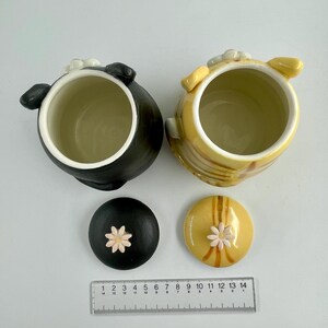 May include: Two ceramic jars with lids. One jar is black, the other is yellow with brown stripes. Both have white interiors and small flower accents on the lids. A ruler is placed below the jars, measuring up to 35 cm.