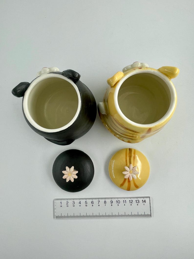 May include: Two ceramic jars with lids. One jar is black, the other is yellow with brown stripes. Both have white interiors and small flower accents on the lids. A ruler is placed below the jars, measuring up to 35 cm.