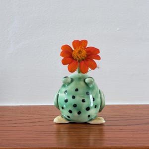 May include: A light green ceramic frog-shaped vase with dark green spots and two small protrusions on top. A single vibrant orange flower with a yellow centre is placed inside the opening at the top of the frog's head, creating a decorative display.
