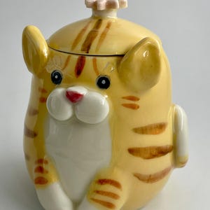 May include: A ceramic cat-shaped jar with a yellow and orange striped pattern. The jar features a white belly, a red nose, and a flower-shaped knob on the lid. The cat has black eyes and white paws, with a cute design.