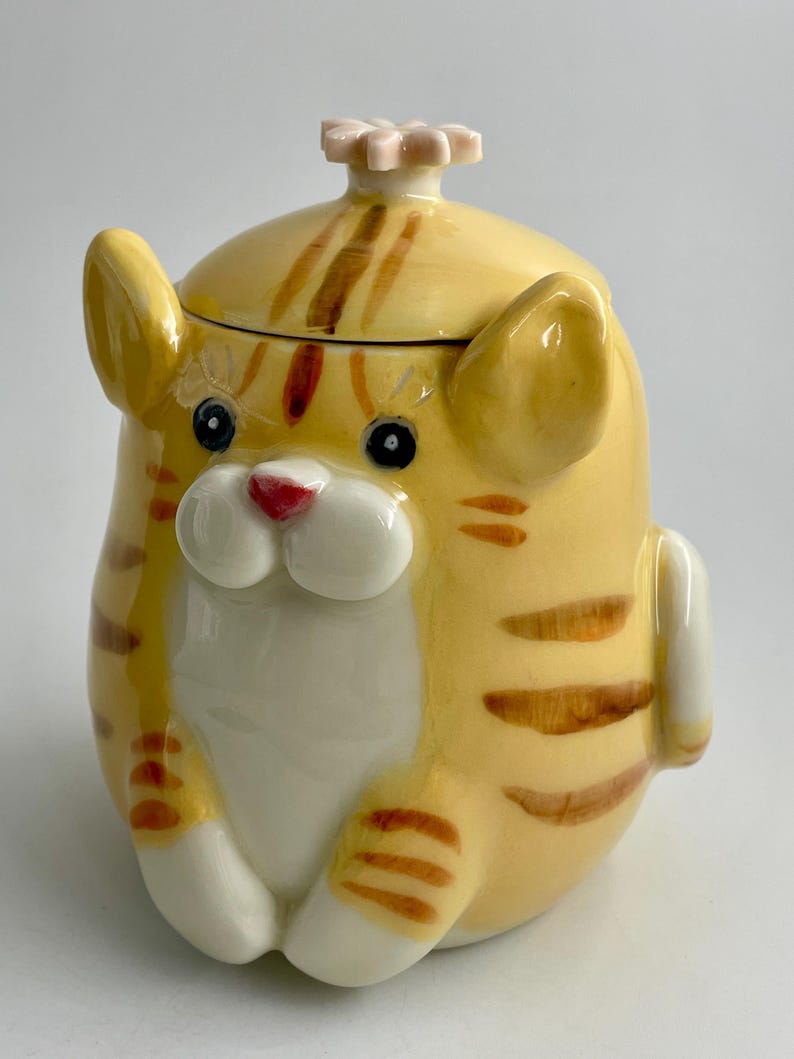 May include: A ceramic cat-shaped jar with a yellow and orange striped pattern. The jar features a white belly, a red nose, and a flower-shaped knob on the lid. The cat has black eyes and white paws, with a cute design.