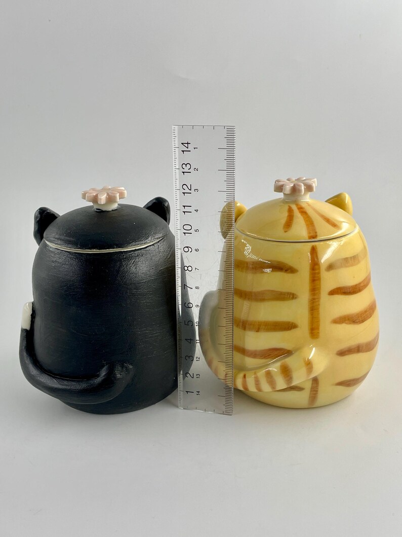 May include: Two ceramic cat-shaped containers. One is black, the other is yellow with brown stripes. Both have lids with a flower-shaped knob. The containers are approximately 13 cm tall, with a tail and ears.