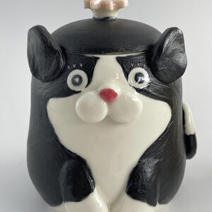 May include: A ceramic cat-shaped jar with a black and white colour scheme. The jar features large, round eyes, a red nose, and a small flower-shaped knob on the lid. This decorative jar is a whimsical item.