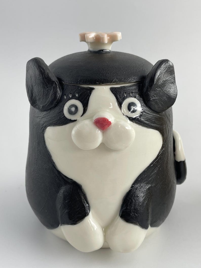 May include: A ceramic cat-shaped jar with a black and white colour scheme. The jar features large, round eyes, a red nose, and a small flower-shaped knob on the lid. This decorative jar is a whimsical item.