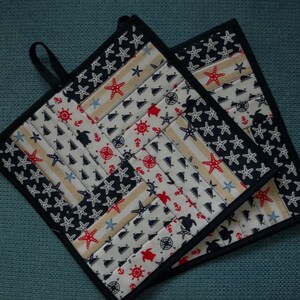 Peut inclure: Deux maniques en tissu noir et blanc sur le thème nautique. Les maniques présentent un motif d'étoiles de mer rouges, d'ancres et de tortues de mer sur un fond blanc avec des rayures bleues. Les maniques ont une bordure noire.