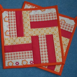 Op de afbeelding: Twee oranje en rode gequilte ovenwanten met een geometrisch patroon. De ovenwanten hebben een lusvormig handvat aan elke hoek.