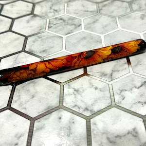 May include: A black pen with a sunflower design. The pen has a black barrel with a silver clip and a black tip. The sunflower design is in shades of yellow, orange, and brown.