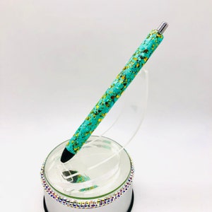 May include: A turquoise and gold glitter pen with a silver clip. The pen is on a clear stand with a silver base.
